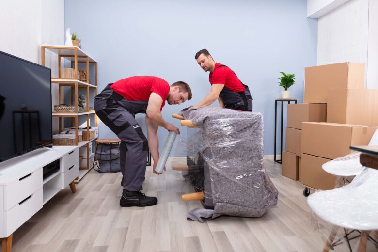 Two Young Male Movers Wrapping The Sofa With Plastic Wrap In Living Room