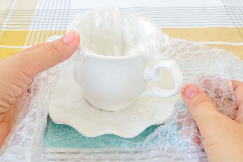 Woman hands packaging white elegant porcelain tea cup and saucer with transparent bubble wrap on a checkered tablecloth. Material for packing fragile items for safe transportation. Close-up.